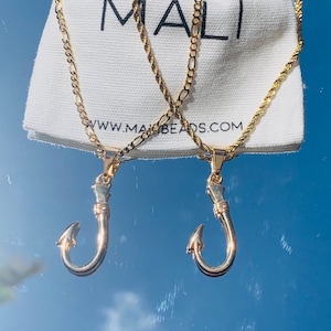 May include: Two gold necklaces with a fish hook pendant. The necklaces are on a white fabric bag with the text "MALI" and "WWW.MALIBEADS.COM".