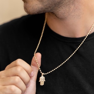 Puede incluir: Un collar de cadena de oro con un colgante de mano de hamsa. La cadena es de estilo cadena de bola.