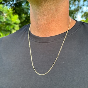May include: A gold chain necklace with a delicate, fine link design. The chain is worn by a person with a black shirt.