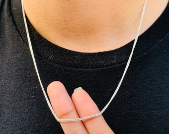 May include: A silver chain necklace with a delicate, flat curb link design. The necklace is displayed against a black shirt, highlighting its bright, metallic appearance. The chain is held by a person's fingers.