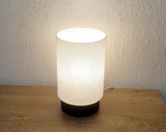 Op de afbeelding: Een witte tafellamp met een getextureerde cilindrische kap en een zwarte voet. De lamp is aan en geeft een warm wit licht.