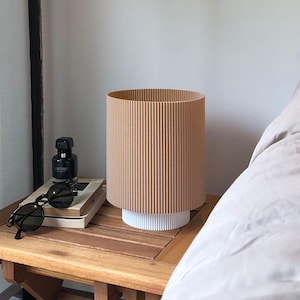 Puede incluir: Una mesita de noche con una tapa de madera marrón y una lámpara blanca con una pantalla corrugada beige. La lámpara está sobre la mesa junto a un libro, gafas de sol y una pequeña botella de perfume.