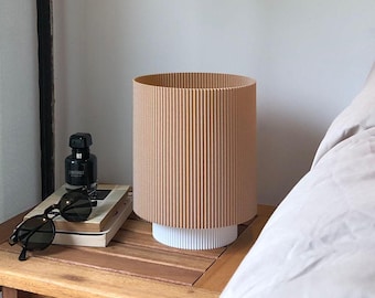 Puede incluir: Una mesita de noche con una tapa de madera marrón y una lámpara blanca con una pantalla corrugada beige. La lámpara está sobre la mesa junto a un libro, gafas de sol y una pequeña botella de perfume.