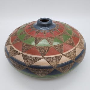 Op de afbeelding: Een handgemaakte keramische vaas met een uniek geometrisch ontwerp. De vaas heeft een afgeronde vorm met een kleine opening. Het oppervlak is versierd met driehoeken in rood, groen en beige, omlijnd met bruine lijnen. Een blauwe band omcirkelt de basis.