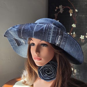 May include: A denim sun hat with a wide brim and a matching denim rose accent. The hat is made from blue denim fabric and has a stylish, casual look. The hat is displayed on a mannequin head.