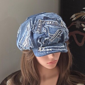 May include: A blue denim newsboy cap featuring a patchwork design with frayed edges, visible white topstitching, and metallic stud accents. The cap is constructed from multiple layered denim fabric pieces with a structured brim and a button detail.