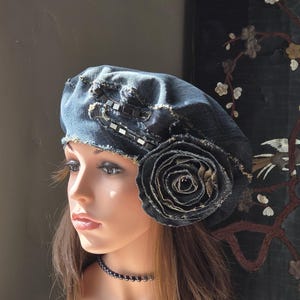 May include: A dark blue denim beret featuring a large, layered fabric rose embellishment on the side. The hat has frayed edges and a decorative strip of small, square, dark metallic studs along the upper section, creating a textured appearance.