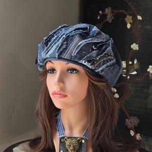 May include: A blue denim beret featuring frayed edges, layered fabric strips, and metallic stud accents. Below the beret, a dark denim collar piece is adorned with a central brass-toned leopard head emblem secured by two metallic rivets.
