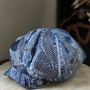 Puede incluir: Una gorra de estilo boina confeccionada con varios parches de tela vaquera en tonos azules. La gorra presenta bordes deshilachados visibles y patrones de costura blanca en los paneles, con un pequeño botón metálico en el lateral de la visera.