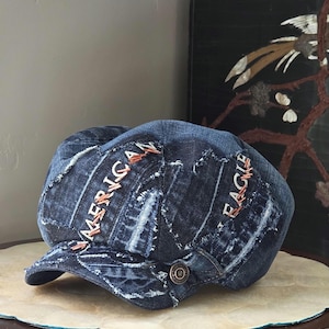 May include: A dark blue denim newsboy cap featuring a patchwork design with frayed edges. The word AMERICAN is stitched in copper-colored thread on the left side, and EAGLE is stitched in the same thread on the right, with a metal button on the side.