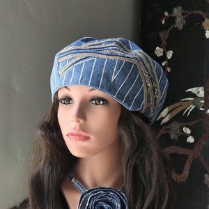May include: A blue denim beret featuring white pinstripes, decorative tan stitching, and metallic studs. The hat is paired with a matching denim choker necklace that displays a large, layered fabric rose made from frayed denim material.