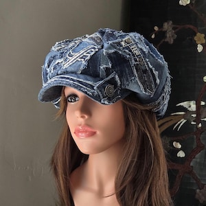 Può includere: Un berretto newsboy in denim blu con un design patchwork, bordi sfrangiati, cuciture a vista e bottoni metallici. Il cappello è realizzato con diverse tonalità di tessuto denim blu, dotato di una visiera strutturata e corona.