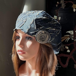Op de afbeelding: Een donkerblauwe spijkerstof baret met een patchworkontwerp, gerafelde randen en metalen kraalversieringen. Een rond accentstuk gemaakt van opgerolde metalen ritssluiting-tanden is aan de zijkant van de hoed bevestigd voor extra textuur.