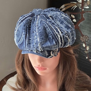 May include: A blue denim newsboy cap featuring a multi-paneled construction with frayed, distressed seams throughout. The hat includes a short, stiff brim and a small silver-toned metal embellishment attached to the side of the cap.