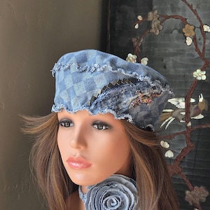 May include: A blue denim beret featuring a checkered pattern, frayed edges, a row of small square metal studs, and a decorative insect-shaped brooch. A matching denim fabric flower choker is worn around the neck of the mannequin head.