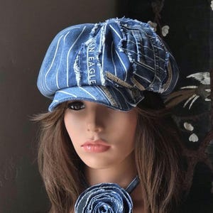 May include: A blue denim newsboy cap featuring white pinstripes, frayed edges, and the text AMERICAN EAGLE printed on a vertical strip. The cap is paired with a matching denim fabric flower accessory worn around the neck on a thin denim band.
