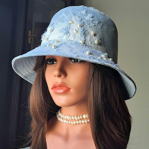 May include: A light blue denim bucket hat with a frayed design and faux pearl embellishments. The hat features a wide brim and is decorated with multiple pearls. The hat is a fashion accessory.