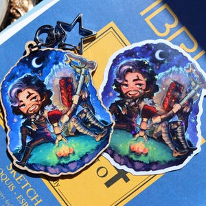 May include: A keychain and sticker set with a cartoon illustration of a character by a campfire under a starry night. The character has a beard, holds a weapon, and wears armour. The keychain has a star-shaped clasp and a silver finish.