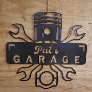 Könnte beinhalten: Schwarzes Garagen-Schild aus Metall mit einem Kolben und Schraubenschlüsseln. Das Schild trägt die Aufschrift "Pat's Garage".