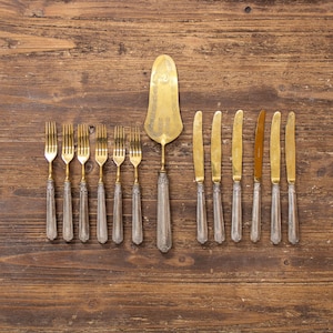 Può includere: Una collezione di posate antiche, tra cui forchette, coltelli e una paletta per torte, disposte su una superficie di legno. Le posate presentano manici ornati e lame e rebbi dorati. La paletta per torte ha un design decorativo.