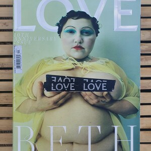 May include: A magazine cover featuring a woman with blue eyeshadow and red lipstick. The cover is titled "LOVE" and features the text "10TH ANNIVERSARY ISSUE" and "BETH". The woman is wearing a yellow robe and has a black and white sticker on her chest that says "LOVE LOVE".