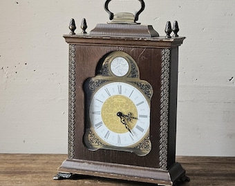 Può includere: Un orologio da camino in legno vintage con un quadrante dorato e indici orari con numeri romani. L'orologio ha un design decorativo attorno al quadrante e un piccolo globo nella parte superiore.