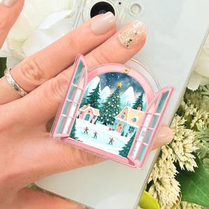 May include: A circular phone grip featuring an illustration of an open window looking out onto a winter landscape with snow-covered houses, pine trees, and figures ice skating on a frozen pond under a starry night sky, framed in pink.