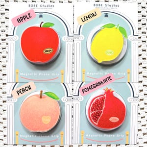 May include: Four magnetic phone grips on display cards, each featuring a fruit design: a red apple labeled FRESH, a yellow lemon labeled ORGANIC, a peach labeled PEACH, and a red pomegranate labeled POMEGRANATE. Each card is marked BOBE Studios.