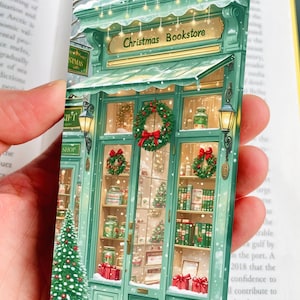 Puede incluir: Ilustración de un marcapáginas de una librería navideña. El marcapáginas presenta una representación detallada de una tienda festiva con una fachada verde, coronas y cajas de regalo. El letrero de la tienda dice "Christmas Bookstore."