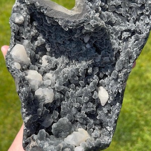 May include: A large, dark gray geode featuring a hollow interior lined with small, rounded, charcoal-colored botryoidal crystal formations and several larger, translucent white crystalline clusters scattered throughout the textured surface.