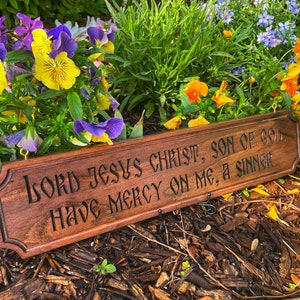 May include: A wooden sign with the text "Lord Jesus Christ, Son of God, Have Mercy on Me, a Sinner" carved into it. The sign is surrounded by purple and yellow flowers.