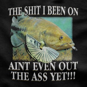 Op de afbeelding: Zwart T-shirt met een vis die een sigaret rookt en geld vasthoudt. De tekst hierboven luidt "THE SHIT I BEEN ON" en hieronder "AIN'T EVEN OUT THE ASS YET!!!"