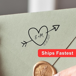 May include: A sage green envelope featuring a black ink stamp of a heart pierced by an arrow with the letters E+M and the date 10.10.2027. A circular gold wax seal with an embossed leaf design is centred on the bottom flap of the envelope.