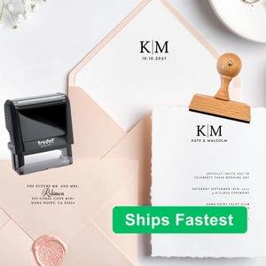 May include: A black Trodat self-inking stamp and a wooden handle rubber stamp featuring the initials KM. The stationery includes a light pink envelope with a wax seal and a white wedding invitation card with deckled edges and black text.