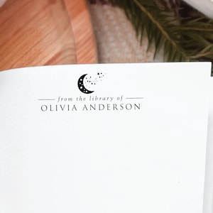 May include: A white book with a black and white crescent moon and stars design. The text "from the library of" and "OLIVIA ANDERSON" is printed below the design.