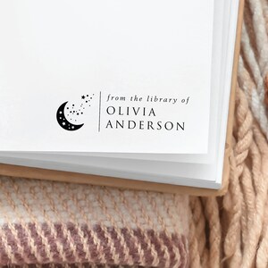 May include: A white book with a black and white stamp that reads "from the library of Olivia Anderson". The stamp features a crescent moon with stars.