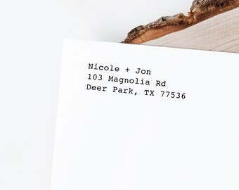 May include: A white rectangular paper featuring black typewriter-style text that reads Nicole + Jon, 103 Magnolia Rd, Deer Park, TX 77536. The paper is positioned partially beneath a piece of natural wood with a rough, textured bark edge.
