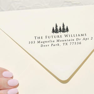 May include: A white envelope with a return address label featuring a black silhouette of a group of pine trees. The text on the label reads "THE FUTURE WILLIAMS 103 Magnolia Mountain Dr Apt 2 Deer Park, TX 77536".