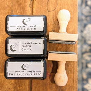 May include: Three black and white self-inking stamps with a crescent moon and stars design. The stamps read "from the library of" followed by the name of the owner. The stamps are on a wooden surface with wooden stamp handles.