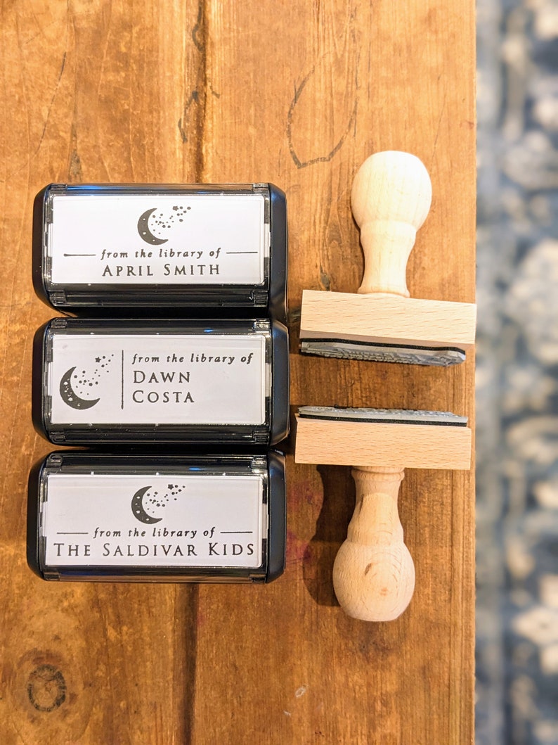 May include: Three black and white self-inking stamps with a crescent moon and stars design. The stamps read "from the library of" followed by the name of the owner. The stamps are on a wooden surface with wooden stamp handles.