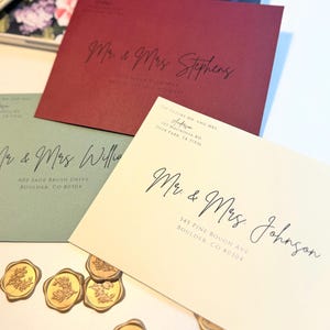 K&ouml;nnte beinhalten: Drei Hochzeitseinladungen mit kalligrafischen Namen und Adressen. Die Umschl&auml;ge sind in Rot-, Gr&uuml;n- und Cremet&ouml;nen gehalten. Die Umschl&auml;ge sind mit goldenen Siegellacken verziert.
