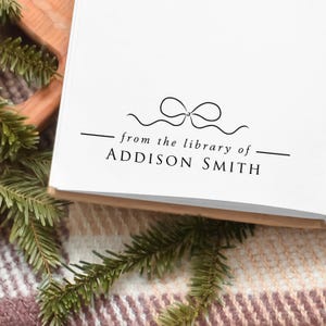 May include: A white book with the text "from the library of" and the name "Addison Smith" in black. A decorative bow graphic is above the text. The book is on a wooden surface with green pine needles.