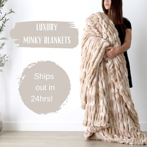 May include: A beige mink blanket with a textured, woven design is held by a person. The blanket is labeled "LUXURY MINKY BLANKETS" in the image.