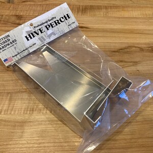 Op de afbeelding: Een zilveren metalen bijenkorf-zitstok, ontworpen en gemaakt in de VS, verpakt in een doorzichtige plastic zak. De verpakking bevat de tekst "Professional Quality Hive Perch Qty 1 Set".