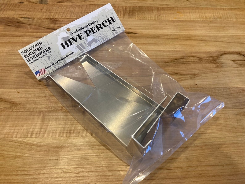 Op de afbeelding: Een zilveren metalen bijenkorf-zitstok, ontworpen en gemaakt in de VS, verpakt in een doorzichtige plastic zak. De verpakking bevat de tekst "Professional Quality Hive Perch Qty 1 Set".