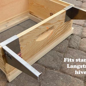 Op de afbeelding: Een houten bijenkorf met twee metalen beugels aan de zijkanten. De kast is gemaakt van licht hout en heeft een gaas aan de bovenkant. De tekst "Fits standard Langstroth hives" staat onder de kast.