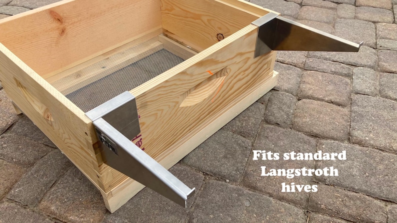 Op de afbeelding: Een houten bijenkorf met twee metalen beugels aan de zijkanten. De kast is gemaakt van licht hout en heeft een gaas aan de bovenkant. De tekst "Fits standard Langstroth hives" staat onder de kast.