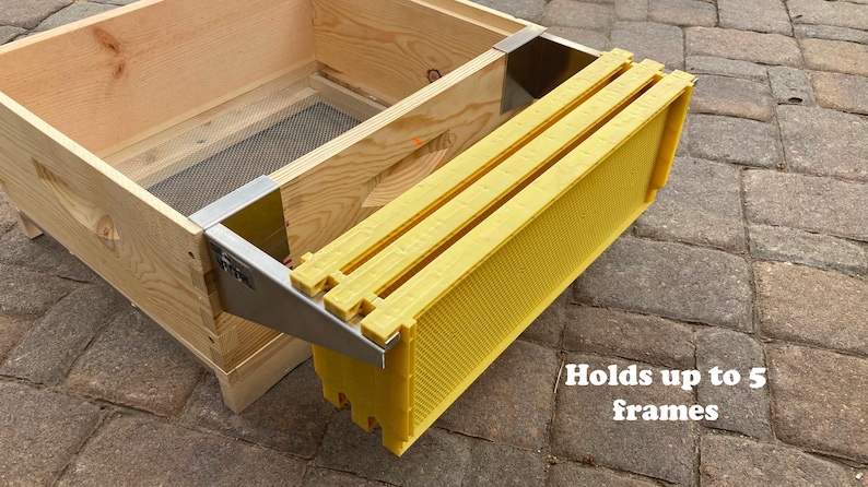Op de afbeelding: Een metalen framehouder voor bijenteelt, bevestigd aan een houten bijenkorf. De houder is ontworpen om maximaal 5 frames te houden en is gemaakt van roestvrij staal. Het is een bijenteeltgereedschap voor het beheren van bijenkorven.