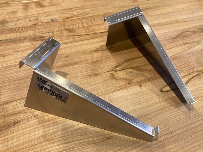 Op de afbeelding: Twee zilveren metalen beugels, een met het label "LEFT SIDE". De beugels zijn schuin en hebben een rechthoekige vorm.