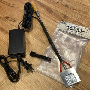 May include: A 115V oxalic acid vaporizer with a black power adapter, a long handle featuring a wooden grip, and a metal heating element. A small black plastic scoop is included, along with a clear plastic bag labeled 115V OXALIC ACID VAPORIZER.